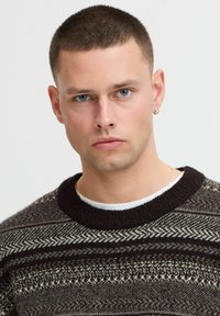 Knit sweater in dark tones featuring geometric patterns and a thick black ribbed crew neckline over a white shirt. Silver earring visible.