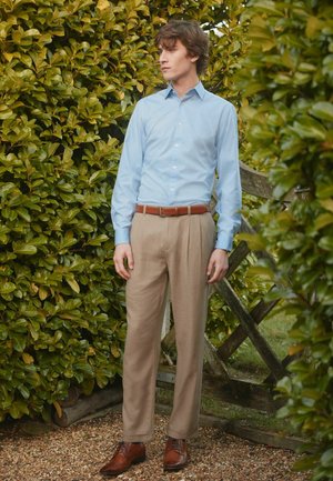 Hellblaues Button-Up-Hemd, beigefarbene, gefaltete Hose, brauner Ledergürtel und passende braune Schuhe. Umgeben von Grünpflanzen und einem Holzzaun.