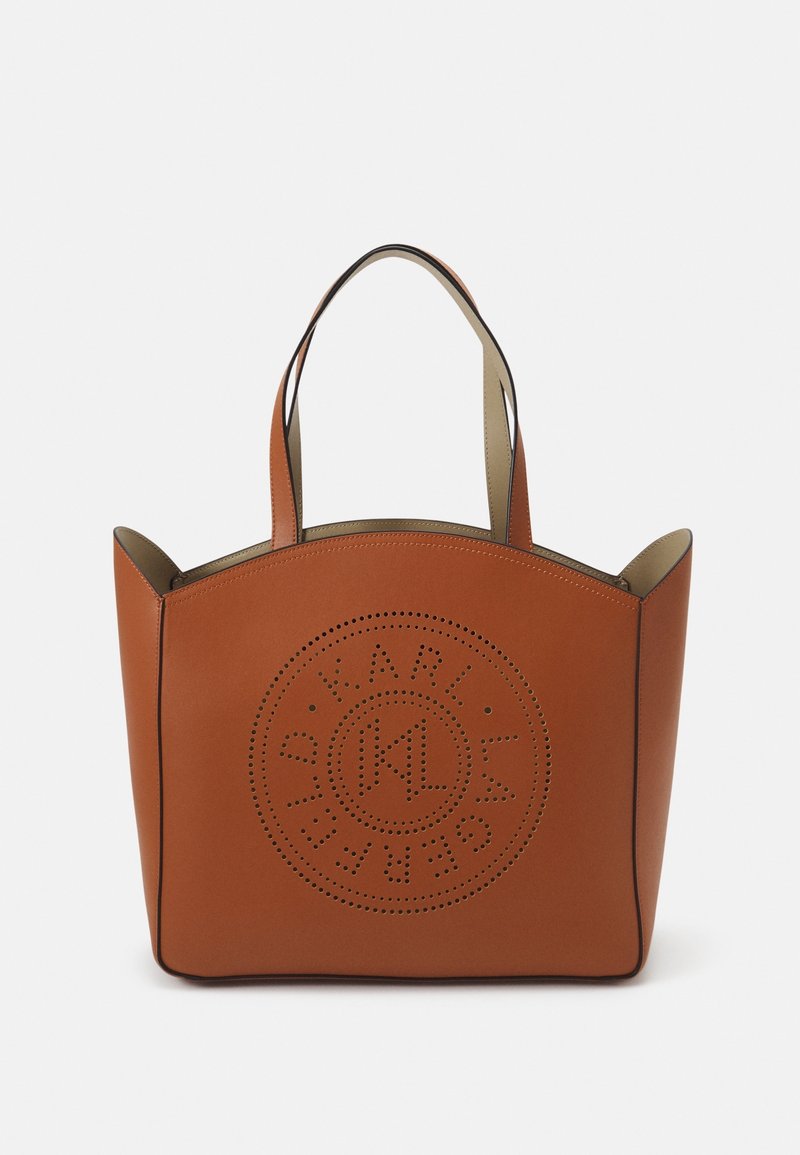 Sac cabas en cuir marron avec deux anses et un logo circulaire perforé sur le devant, sur un fond uni.