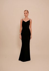 Žena s povučenom ravnom frizurom nosi dugu, usku crnu haljinu s tankim trakama ispred jednostavne bež pozadine.