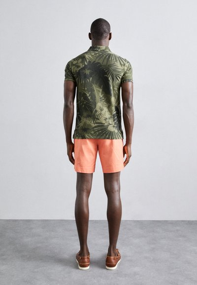 Polo shirt cu model floral verde, pantaloni scurți corali, sandale maro; modelul întors; textură netedă și siluetă ajustată.