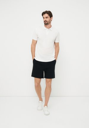 Uomo in piedi con le mani nelle tasche, indossa una polo bianca, pantaloncini neri e sneakers bianche su sfondo bianco neutro.