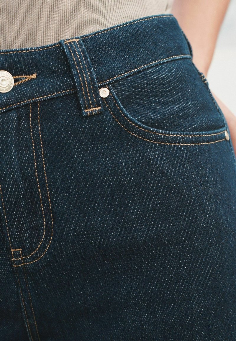 Gros plan d'une personne portant un jean en denim bleu foncé avec des coutures orange et un haut côtelé beige rentré à l'intérieur.