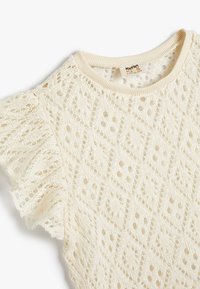 Top in pizzo lavorato a maglia di colore bianco sporco con motivo a diamante, finitura testurizzata e dettagli arricciati sulle spalle. Presenta un collo rotondo e un tessuto morbido.