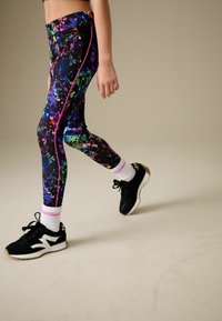 Flerfärgade leggings med en färgstänkdesign, med svart bas och klara accenter, kompletterade med svarta sportskor och vita strumpor.