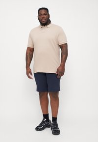 Beige polo skjorte med krave, parret med marineblå shorts. Modellen har sorte atletiske sko med lilla accenter på. Enkel, minimalistisk design.