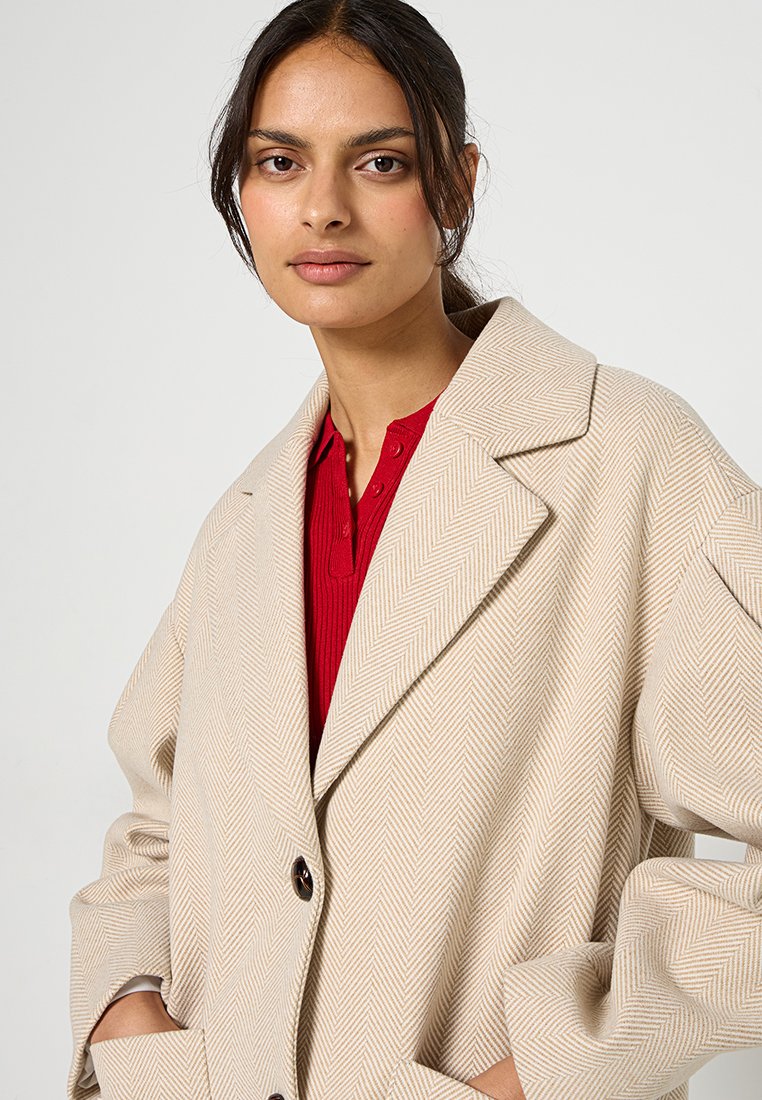 Beżowy płaszcz w wzór jodełki z wykładanym kołnierzem, o luźnym kroju, z ciemnym zapięciem na guziki, nałożony na czerwony top z prążkami.