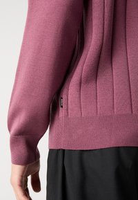 Gros plan sur le bras gauche et le torse d'une personne portant un pull côtelé mauve avec une petite étiquette Boss et un pantalon noir.