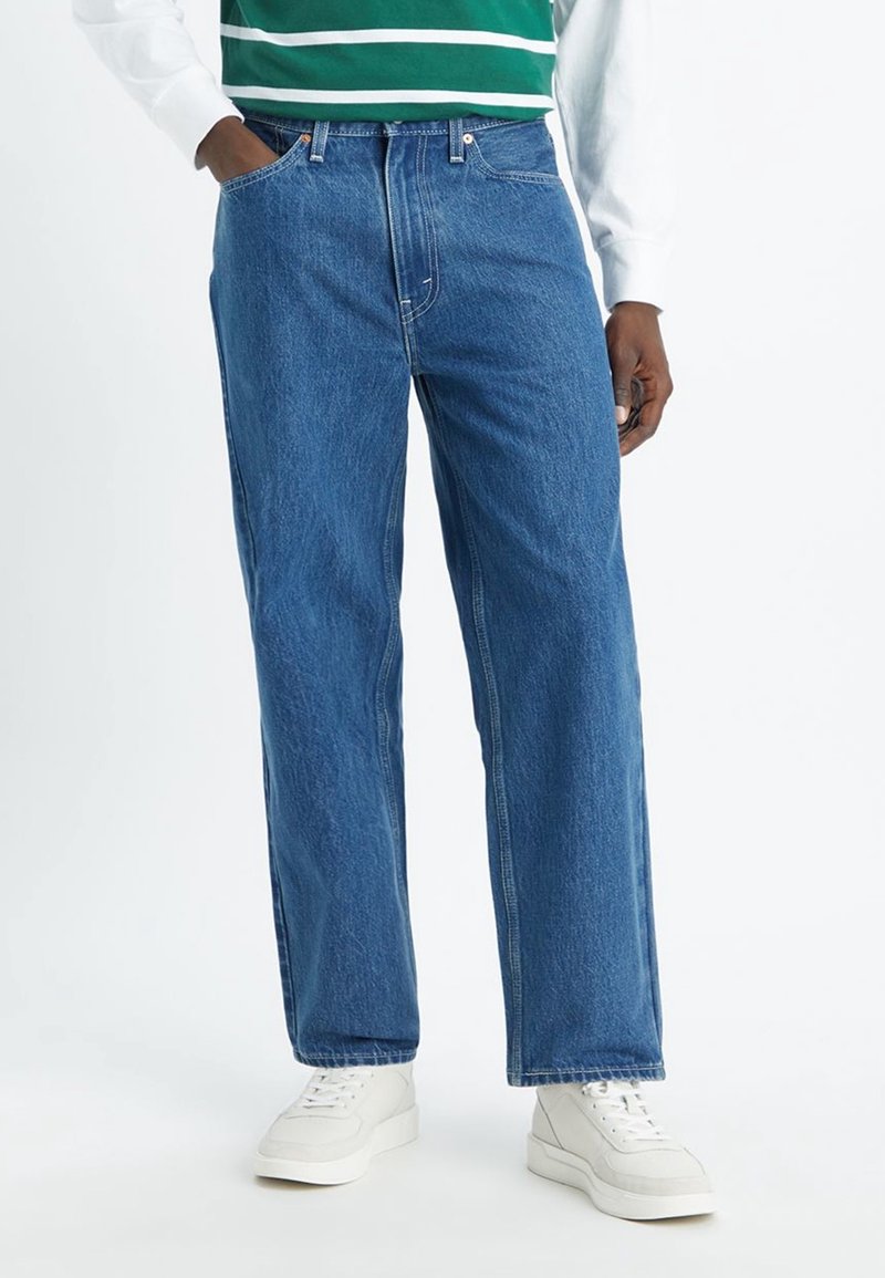Homme portant un jean droit bleu, des baskets blanches et un haut rayé vert et blanc, avec une main dans la poche.