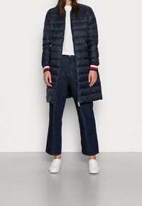 Granatowy puchowy płaszcz o długiej sylwetce, pikowanej fakturze i mankietach w czerwono-białe paski, noszony na ciemnych dżinsach o szerokich nogawkach i białych sneakersach.