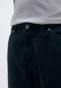 Σκούρο ναυτικό παντελόνι από υφασμάτινη ύφανση, με κουμπί κλεισίματος και μεταλλικό λογότυπο, καθώς και γωνιακές τσέπες. Φορεμένο με ανοιχτό γκρι μπλουζάκι.