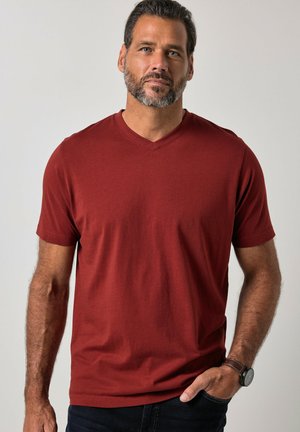 Mann mit Bart, der ein rotes kurzärmeliges V-Neck-T-Shirt und eine Armbanduhr trägt, steht mit einer Hand in der Tasche vor einem einfachen Hintergrund.