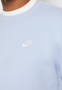 Vaaleansininen collegepaita, jossa on valkoinen pyöreä kaula-aukko ja valkoisella brodeerattu Nike-logo rinnassa. Pehmeä puuvillasekoitemateriaali, jossa on sileä rakenne.
