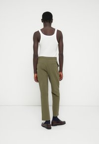 Άνδρας στέκεται με το πρόσωπο στον τοίχο, φοράει λευκό αμάνικο πουκάμισο, χακί παντελόνι, ναυτικό καλσόν και καφέ δερμάτινα παπούτσια.