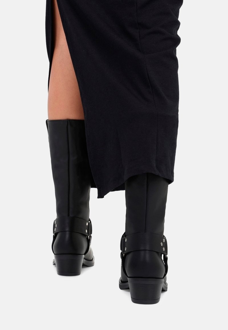 Czarne skórzane kozaki za kolano na słupku z detalami w postaci pasków, noszone z czarną spódnicą z wysokim rozcięciem po boku.