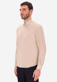 Camicia beige chiaro a maniche lunghe realizzata in tessuto morbido, con colletto, chiusura frontale con bottoni e orlo leggermente curvo.