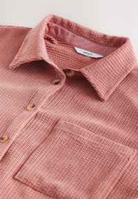 Camicia in velluto a coste rosa con superficie strutturata, chiusura con bottoni e una tasca sul petto. Presenta un colletto piegato e una fodera interna a contrasto.