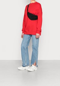 Czerwona, oversize'owa bluza z długim rękawem, jasnoniebieskie dżinsy z bocznymi rozcięciami oraz białe sneakersy, dopełnione czarną torebką na ramię.