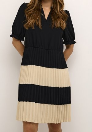 Femme portant une robe courte jusqu'aux genoux, à manches courtes, avec des rayures horizontales plissées noires et beiges, debout, les mains dans le dos.