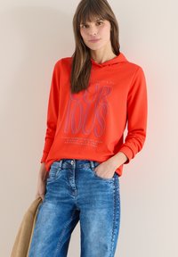 Femme portant un sweat à capuche orange vif avec le texte "CURIOUS", un jean bleu, tenant une veste beige, debout contre un mur clair uni.