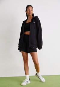 Juodas užtrauktuku užsegamas džemperis su dideliais priekiniais kišenėmis, derinamas su juodu trumpu marškinėliu ir šortais. Batai su baltais ir juodais akcentais užbaigia aprangą.