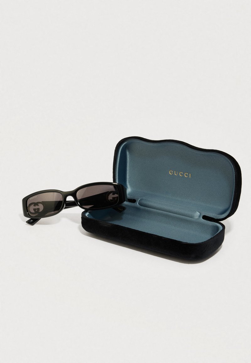 Schwarze, rechteckige Sonnenbrille mit dunklen Gläsern und Logo, in einem weichen schwarzen Samt-Etui mit türkisfarbenem Innenfutter.