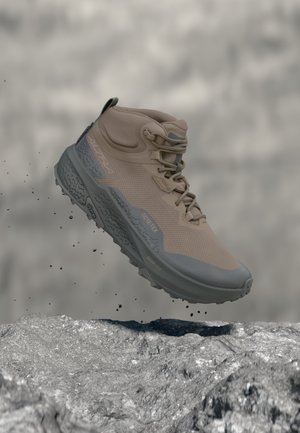 Ανοιχτό καφέ πεζοπορικό μποτάκι με γκρι σόλα και λογότυπο Gore-Tex εμφανίζεται στον αέρα πάνω από βραχώδες έδαφος με πτώση σωματιδίων χώματος.