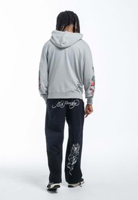 Sudadera con capucha gris con diseños florales en las mangas, combinada con pantalones negros que tienen acentos gráficos en blanco. Ajuste relajado, estilo casual.
