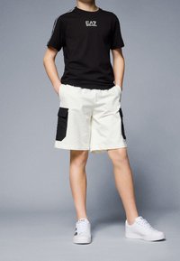 Černé tričko s logem EA7, bílé šortky s černými kapsami na bocích, bílé tenisky, hladké materiály, volný střih a kontrast barev.