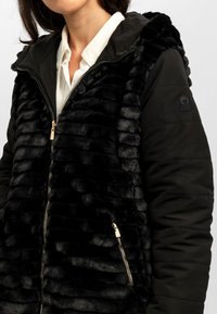 Zwarte geïsoleerde jas met een pluche nepbont voorkant en een ritsluiting, en een gladde stof achterkant. Versterkt met een capuchon en gouden hardware.