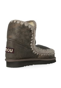 Stivale alla caviglia in suede marrone con cuciture beige, fodera morbida, suola piatta e una patch con logo laterale. Presenta una punta arrotondata e dettagli texture.