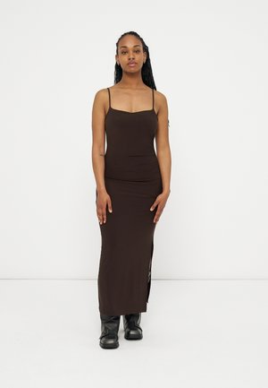 Kvinde stående, iført en tætsiddende mørkebrun ærmeløs kjole med sideslids og sorte læderstøvler mod en ensfarvet hvid baggrund.