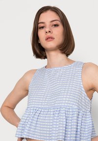 Top sem mangas em padrão xadrez gingham azul claro e branco com bainha peplum franzida. Feito de um tecido leve, apresentando um decote redondo.