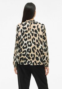 Woman with long black hair wearing a beige and black leopard print blouse and black pants, shown from the back against a white background.