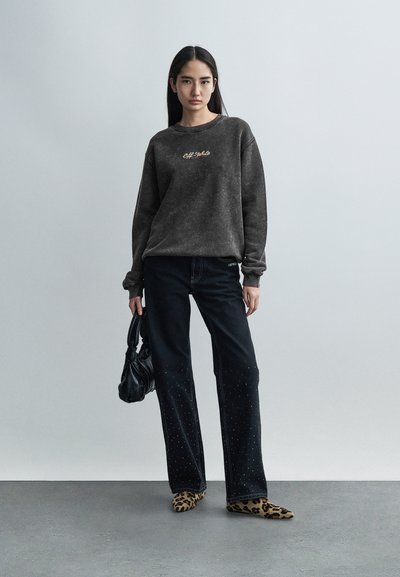 Σκούρο γκρι φούτερ με χρυσό λογότυπο, συνδυασμένο με μαύρα τζιν με διακοσμητικές πινέζες. Το μοντέλο φοράει παπούτσια με λεοπάρ μοτίβο και κρατάει μια μαύρη τσάντα.