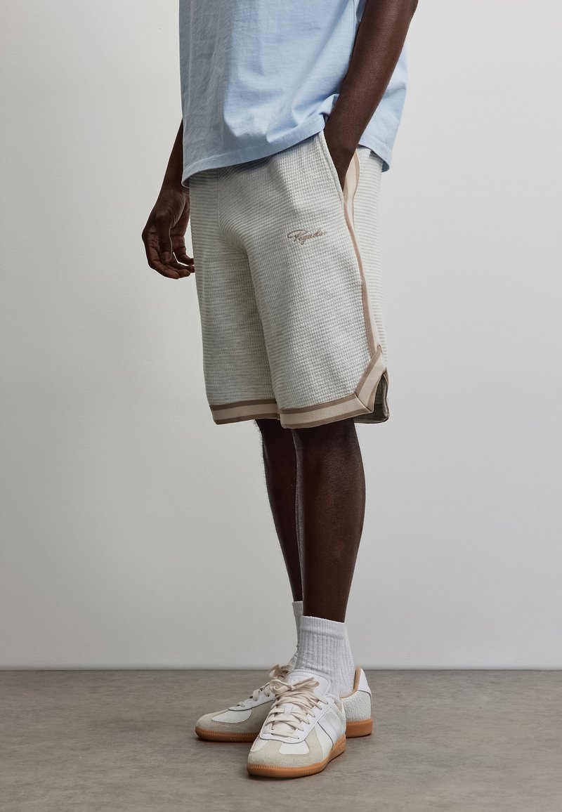 Homme portant une chemise bleu clair, un short beige texturé avec une bordure marron, des chaussettes blanches et des baskets beiges, debout avec une main dans la poche.