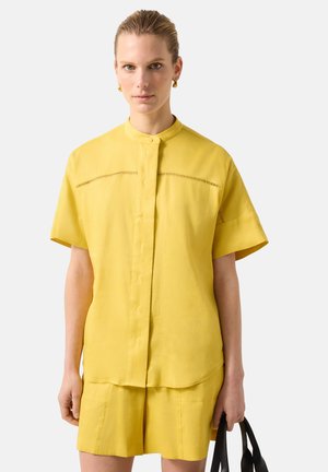 Kobieta ubrana w żółtą koszulę na guziki z krótkim rękawem i pasujące do niej szorty, trzyma czarną torebkę w prawej ręce.