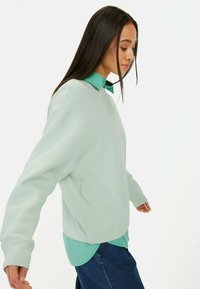 Licht muntgroene gebreide sweatshirt met verlaagde schouders, gelaagd over een muntgroene blouse, gecombineerd met donkerblauwe jeans.