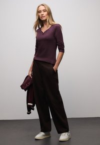 Top manica lunga di colore viola intenso con scollo a V; abbinato a pantaloni larghi marrone scuro e scarpe da ginnastica bianche; tiene in mano una giacca bordeaux abbinata.