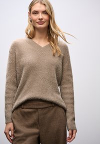 Beige, strukturierter V-Ausschnitt-Pullover aus weichem, fuzzeligem Material; getragen über braunen, taillierten Hosen mit geradem Schnitt und frontalen Taschen.
