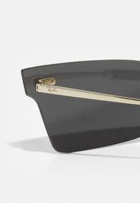 Occhiali da sole con lenti grigie e una montatura in metallo dorato, dotati di una cerniera e logo inciso sul lato. Forma angolare e design elegante.