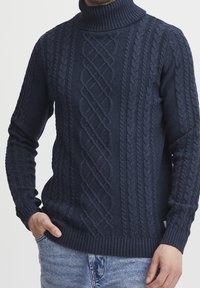 Homme portant un pull col roulé en maille torsadée bleu marine avec des motifs texturés et un jean en denim bleu clair, les mains dans les poches.
