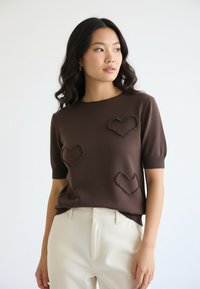 Młoda kobieta o długich ciemnych włosach w brązowym swetrze z krótkim rękawem, ozdobionym fakturowanymi kształtami serc, oraz jasnobeżowych spodniach, stojąca na białym tle.