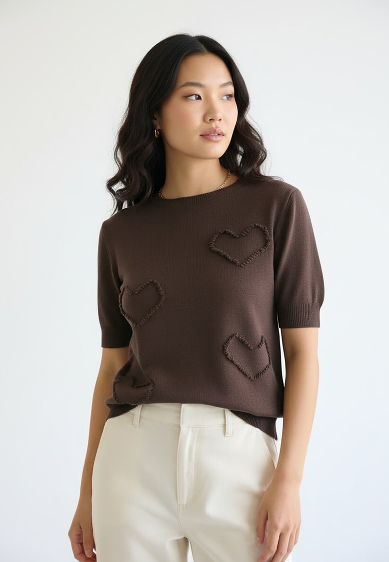 Młoda kobieta o długich ciemnych włosach w brązowym swetrze z krótkim rękawem, ozdobionym fakturowanymi kształtami serc, oraz jasnobeżowych spodniach, stojąca na białym tle.