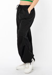 Schwarze Jogginghose mit seitlichen Taschen, lockerer Passform und gerafften Bündchen. Kombiniert mit weißen Turnschuhen, die Schnürsenkel und strukturierte Sohlen haben.