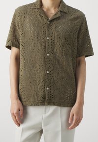 Olijfgroene kortemouwen shirt gemaakt van textuurstof met ingewikkelde uitsnijdingen en een borstzak, voorzien van knopen aan de voorkant.