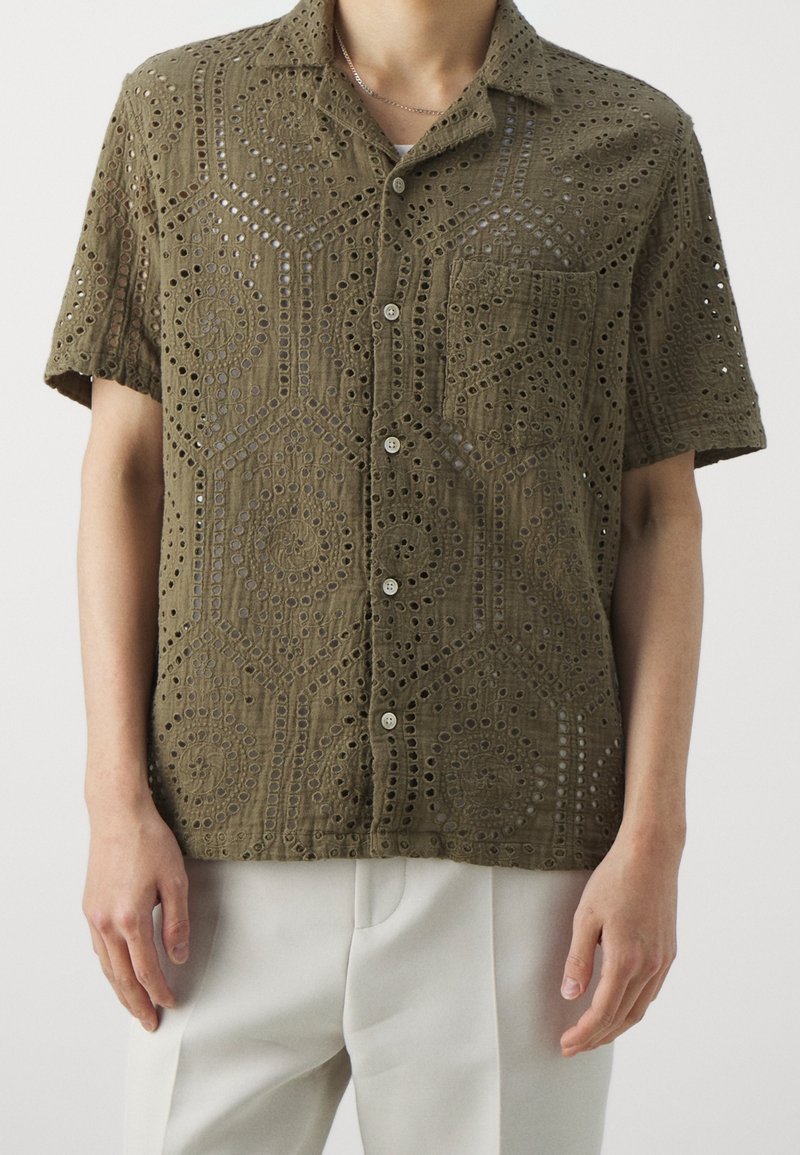 Olijfgroene kortemouwen shirt gemaakt van textuurstof met ingewikkelde uitsnijdingen en een borstzak, voorzien van knopen aan de voorkant.