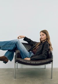 Fauteuil en cuir marron, pieds en chrome ; mannequin portant une veste sombre, un haut marron et un jean évasé bleu clair, des bottines marron, dans une pose détendue.