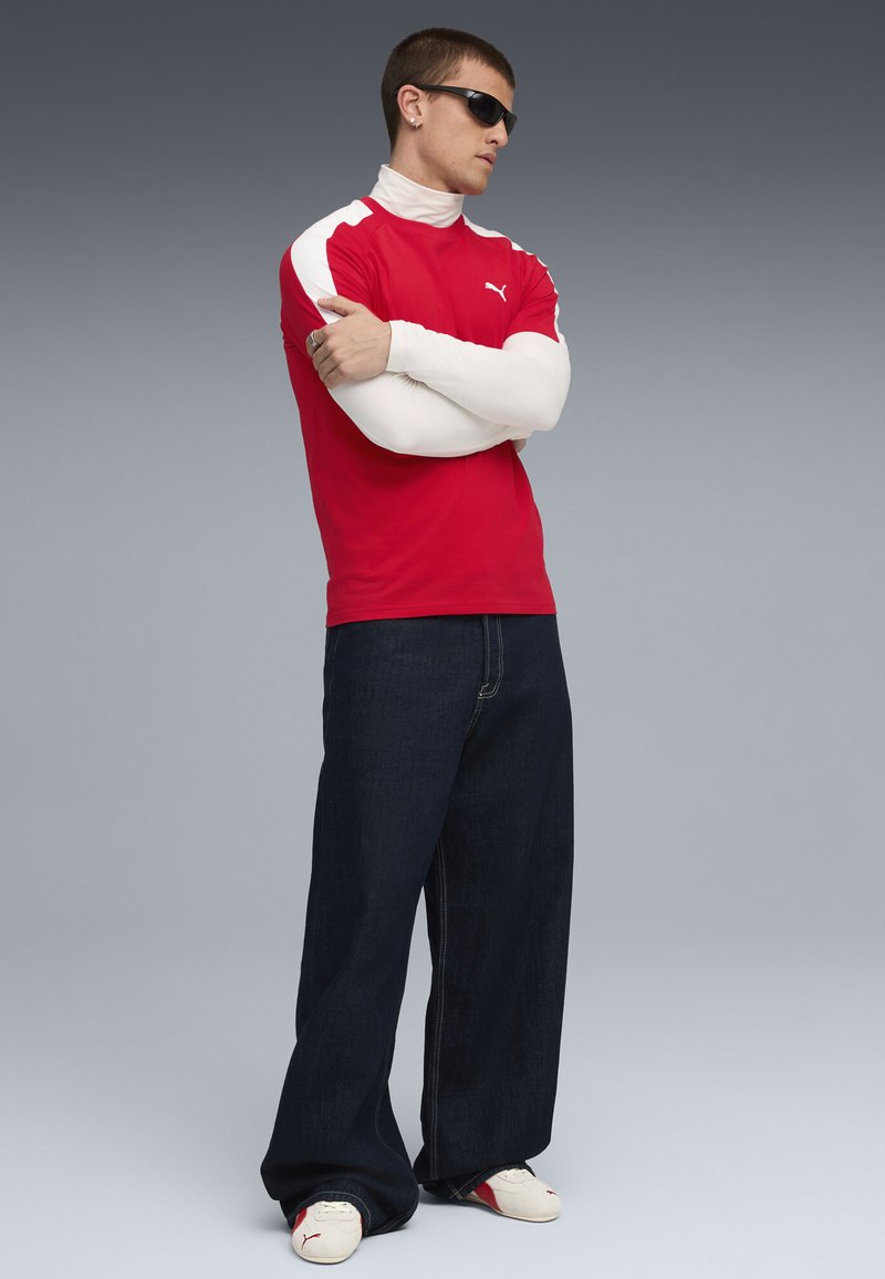 Rød kortærmet skjorte med hvide detaljer, mørkeblå brede bukser og solbriller. Modellen bærer en tætsiddende hvid turtleneck nedenunder.