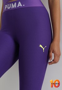 Fioletowe legginsy z prążkowanej tkaniny z białą taśmą z logo "PUMA" w talii oraz żółtym logo PUMA na lewej udzie. Gładka tekstura z dopasowanym krojem.